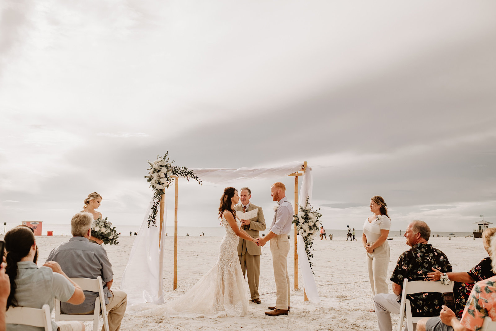 Beach Wedding Ceremony Venue at Hilton Clearwater Beach | White Folding Garden Chairs and Bamboo Arch with Sheer Draping and Greenery Floral Arrangement Tie Backs with White Roses