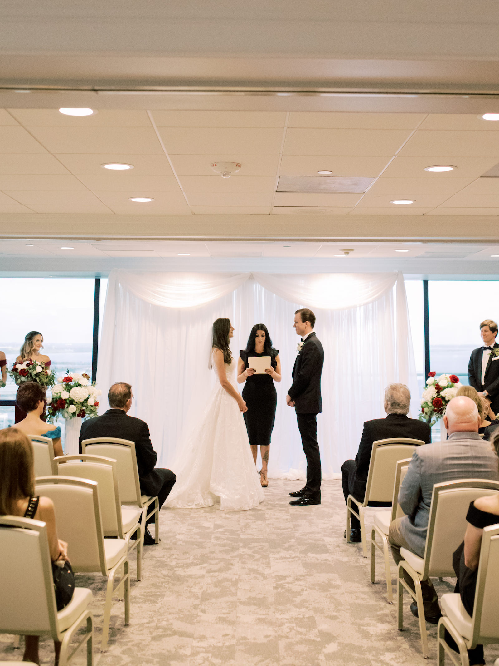 Indoor Tampa Wedding Ceremony at Downtown Tampa Wedding Venue The Tampa Club | Pipe and Drape Ceremony Backdrop | V Neck Embroidered Illusion Panel Allure Couture Designer Wedding Dress Bridal Gown | Groom in Classic Black Suit Tux with Bow Tie