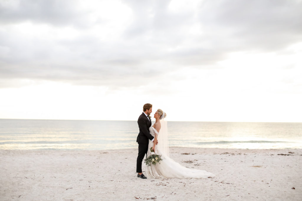 Elegant Sarasota Bride and Groom Walking on Anna Marie Island Beach at Sunset, Wearing Vintage Crystal Headpiece with White Fit and Flare Wedding Dress and Off the Shoulder Lace Sleeves, Holding Lush Ivory Floral Bouquet with Greenery, Groom in Classic Black Tuxedo | Florida Wedding Photographer Lifelong Photography Studio | Kelly Kennedy Weddings and Events