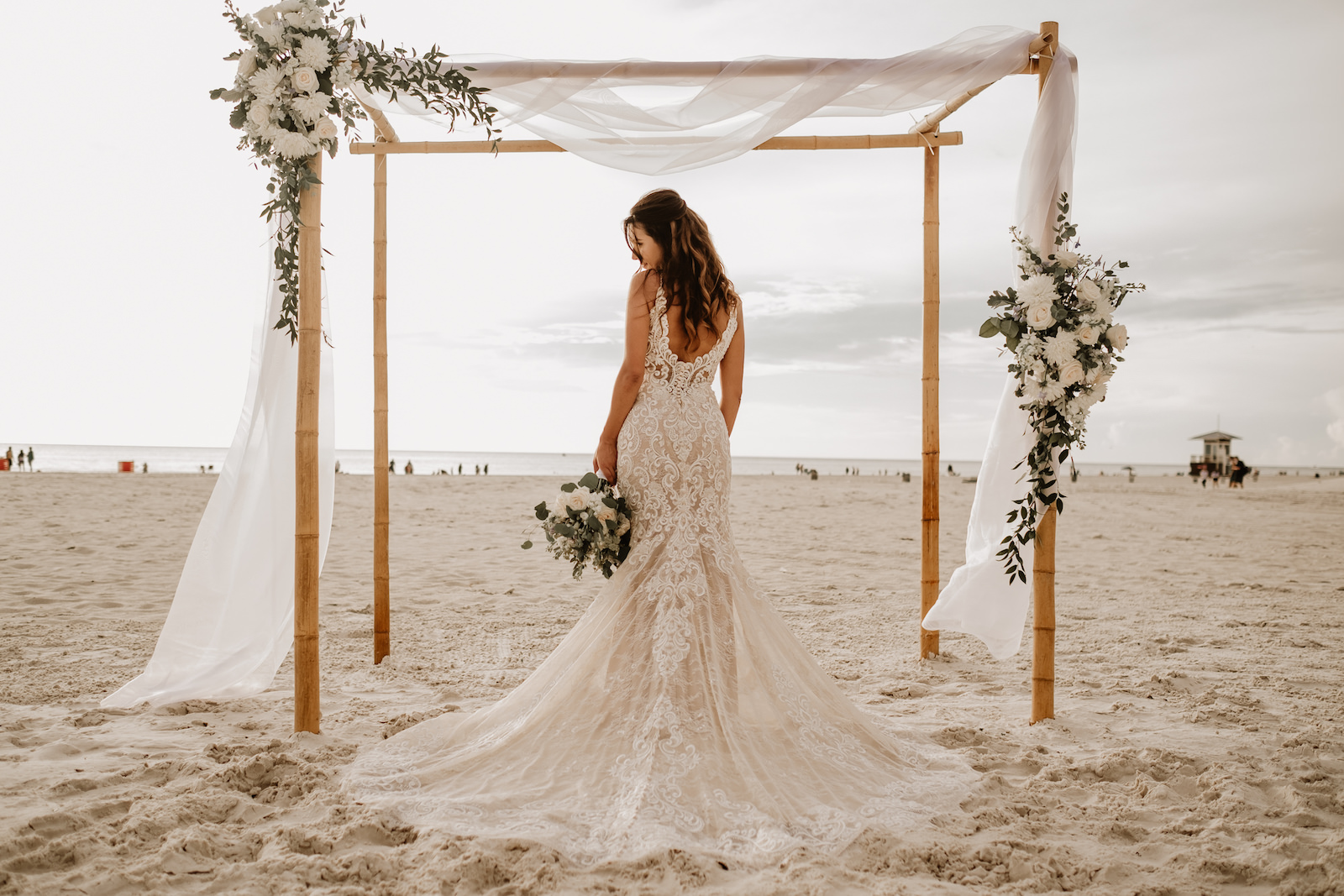 Beach Wedding Ceremony Venue at Hilton Clearwater Beach | White Folding Garden Chairs and Bamboo Arch with Sheer Draping and Greenery Floral Arrangement Tie Backs with White Roses Chrysanthemums and Stock | Allure Bridals Lace Low Back Spaghetti Strap Sheath Bridal Gown Wedding Dress