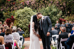 Tampa Bay Bride and Groom Kiss At The End of The Aisle At Sunken Gardens Wedding Ceremony, Bride Wearing Long White BHLDN Wedding Dress with Illusion Lace Sleeves | Florida Wedding Planner John Campbell Weddings