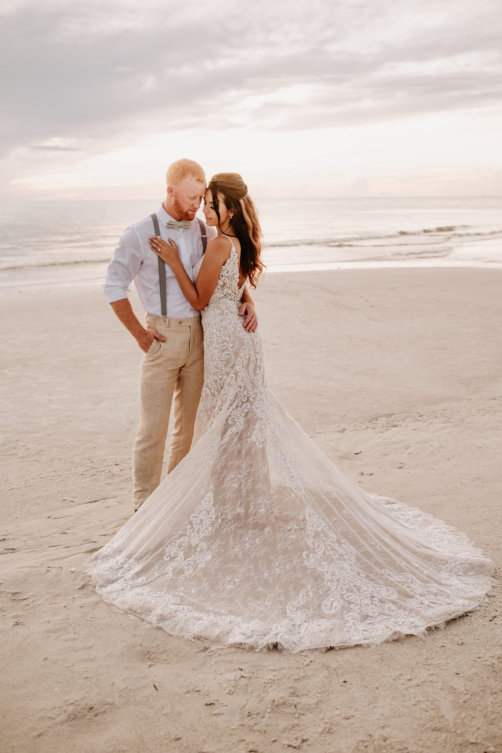 Outdoor Bride and Groom Sunset Portrait at Wedding Venue Hilton Clearwater Beach | Allure Bridals Lace Low Back Spaghetti Strap Sheath Bridal Gown Wedding Dress | Groom Wearing Khaki Pants and Grey Suspenders with White Shirt and Bow Tie