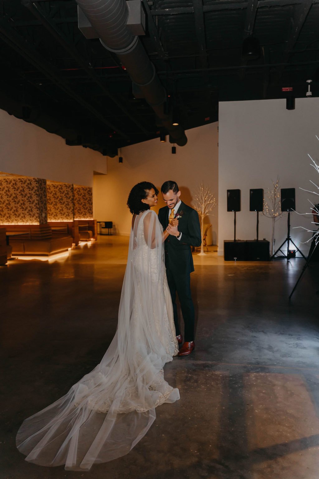 Indoor Bride and Groom First Dance in Tampa Wedding Venue Hyde House | Illusion Lace Embroidered Beaded V Neck Wedding Dress Bridal Gown with Sheer Tulle Cape Sleeves by Designer Amalia Carrara Bridal | Groom in Classic Black Tux Suit with Gold Tie