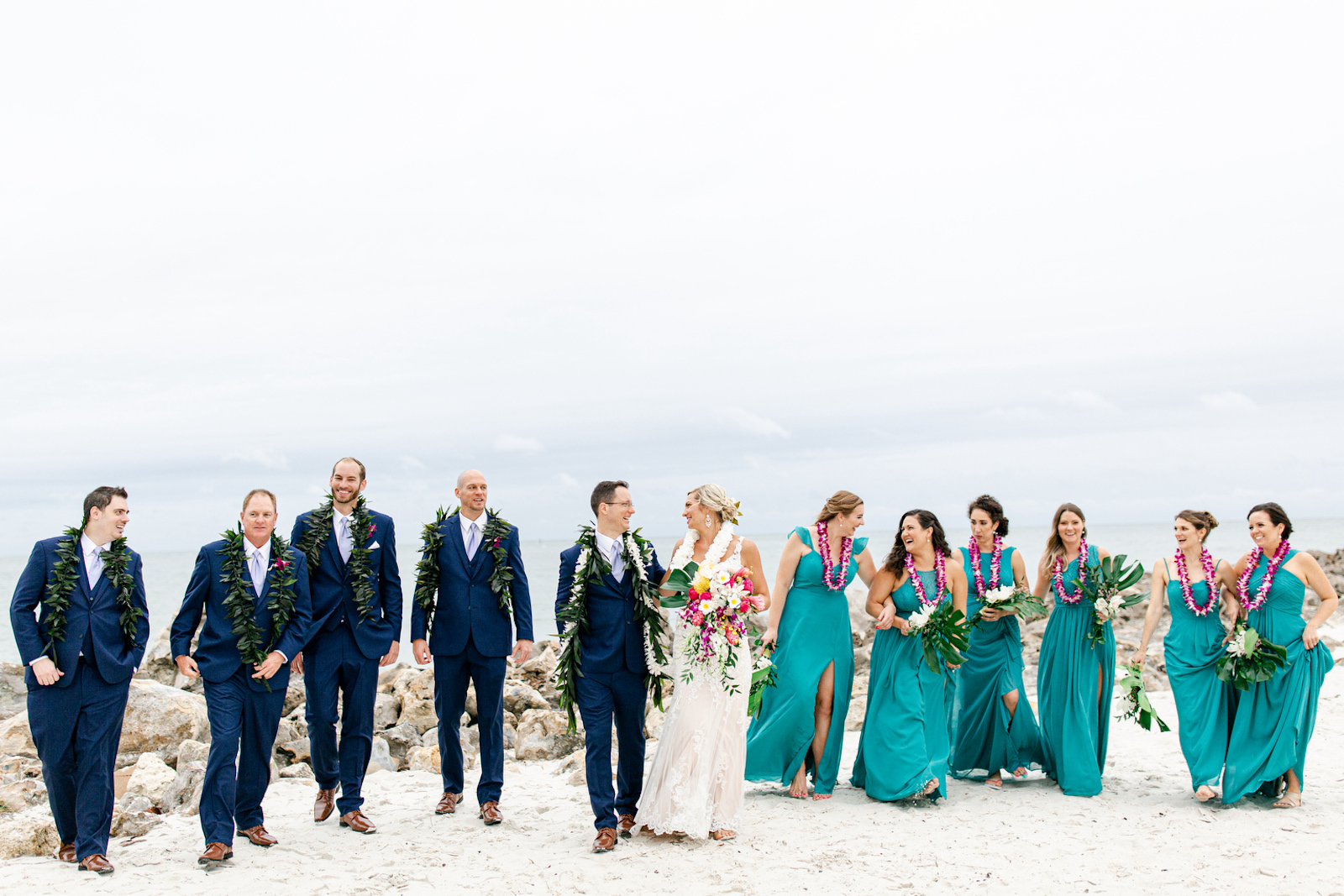 Tropical Elegant Wedding Party Photo on Clearwater Beach, Groomsmen in Blue Suits with Hawaiian Green Leaves Leis, Bride Holding Lush Colorful Floral Bouquet Wearing Lace Dress and All White Floral Leis, Bridesmaids Wearing Teal Mix and Match Dresses Wearing Hawaiian Purple Flower Leis | Tampa Bay Wedding Planner Special Moments Event Planning