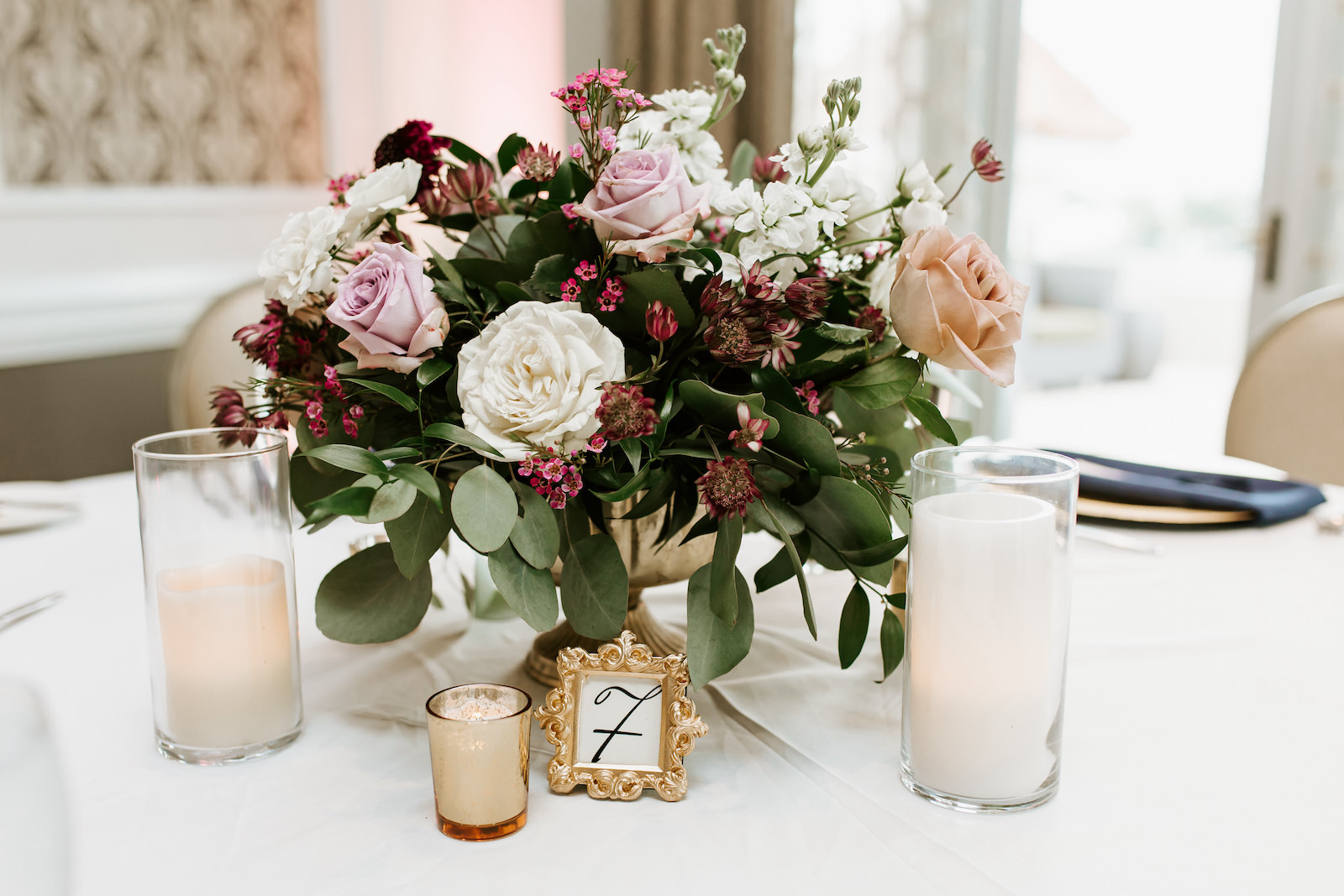 Wedding Reception Tables with Gold Chargers and Ivory Table Linens and Navy Blue Napkins | Low Gold Compote Vase Centerpiece with Loose Natural Eucalyptus Greenery and Ivory and Pink Roses and Candles | Ornate Gold Frame Table Number