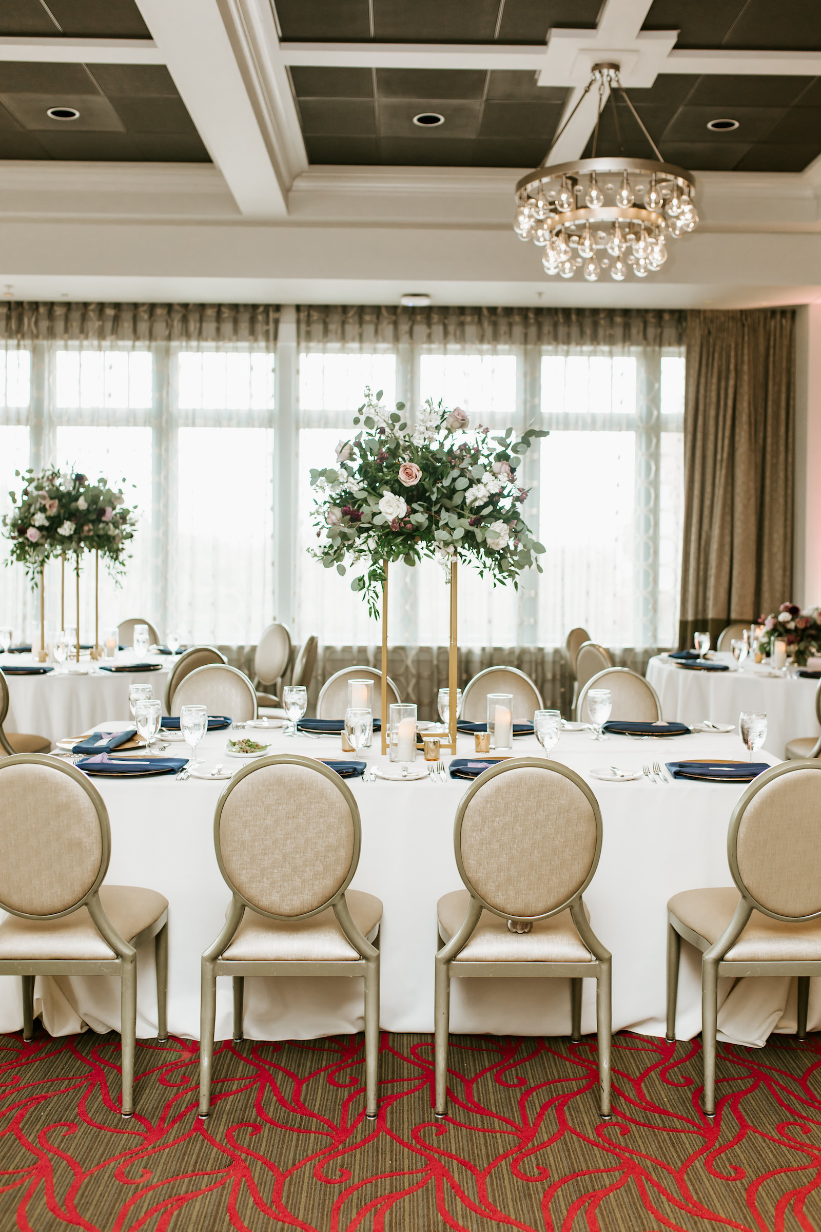 Indoor Hotel Ballroom Reception at St. Pete Wedding Venue The Birchwood | Reception Tables with Gold Banquet Chairs and Ivory Table Linens and Navy Blue Napkins | Tall Gold Metal Centerpiece Stand with Loose Natural Eucalyptus Greenery and Ivory and Pink Roses