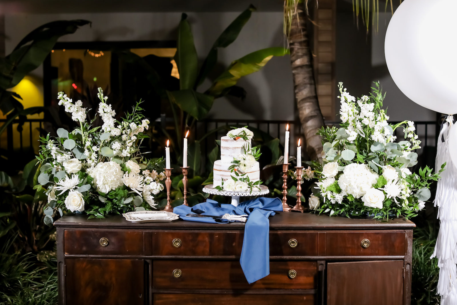 Vintage Inspired Wedding Dessert Table with Two-Tier Semi Naked Iced Wedding Cake with White Frosting, Ivory Florals, and Greenery, Blue Linen Decor with Brass Candlesticks and Lush Tropical Floral Arrangements | Florida Wedding Photographer Lifelong Wedding Photography Studio | Sarasota Wedding Planner Kelly Kennedy Weddings