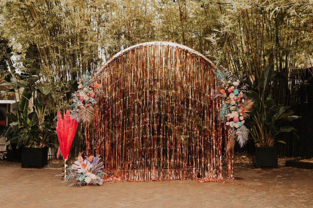 Iridescent Pastel 90's Wedding Inspiration | Wedding Arch Backdrop with Rose Gold Tinsel Curtain and Pastel Flower Arrangements with Painted Leaves