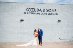 St. Petersburg Bride and Groom at Kozuba & Sons Craft Distillery, Modern Boho Florida Bride Holding Vibrant Autumn-Inspired Floral Bouquet with Orange, Yellow, Red and White Florals, Bride Wearing White Lace Mermaid Style Dress, Groom in Dark Blue Suit | Tampa Bay Wedding Planner Coastal Coordinating