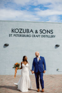 St. Petersburg Bride and Groom at Kozuba & Sons Craft Distillery, Modern Boho Florida Bride Holding Vibrant Autumn-Inspired Floral Bouquet with Orange, Yellow, Red and White Florals, Bride Wearing White Lace Mermaid Style Dress, Groom in Dark Blue Suit | Tampa Bay Wedding Planner Coastal Coordinating