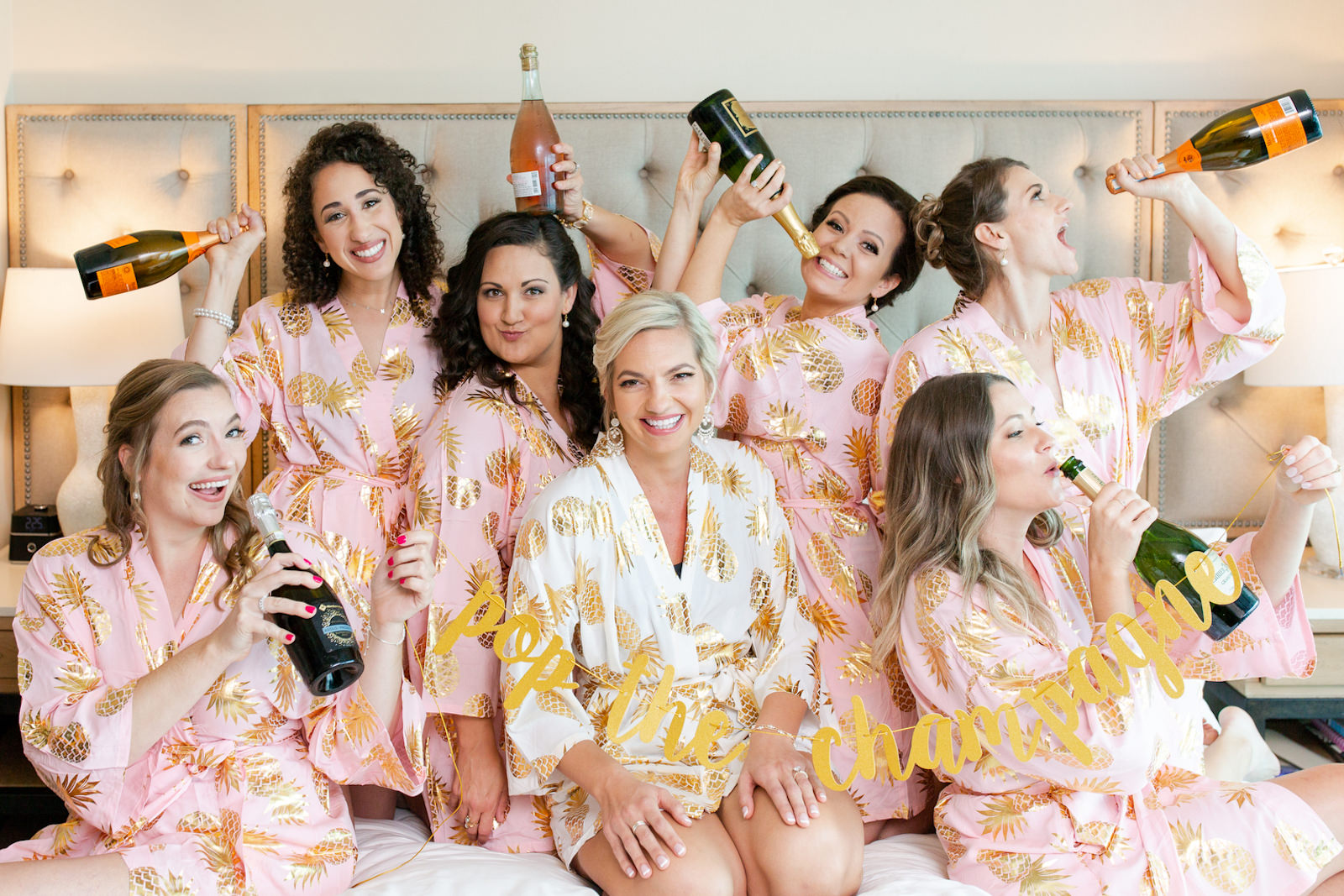 Bride Getting Ready Wedding Photo Wearing White and Gold Foil Pineapple Tropical Robe, Bridesmaids in Matching Pink with Gold Foil Pineapple Robes Drinking Champagne with Pop the Champagne Banner | Special Moments Event Planning