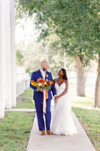 Downtown St. Petersburg Bride and Groom, Modern Boho Florida Bride Wearing White Lace Mermaid Style Dress, Groom in Dark Blue Suit Holding Vibrant Autumn-Inspired Floral Bouquet with Orange, Yellow, Red and White Florals | Tampa Bay Wedding Planner Coastal Coordinating