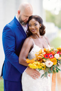 Downtown St. Petersburg Bride and Groom, Modern Boho Florida Bride Holding Vibrant Autumn-Inspired Floral Bouquet with Orange, Yellow, Red and White Florals, Bride Wearing White Lace Mermaid Style Dress, Groom in Dark Blue Suit | Tampa Bay Wedding Planner Coastal Coordinating