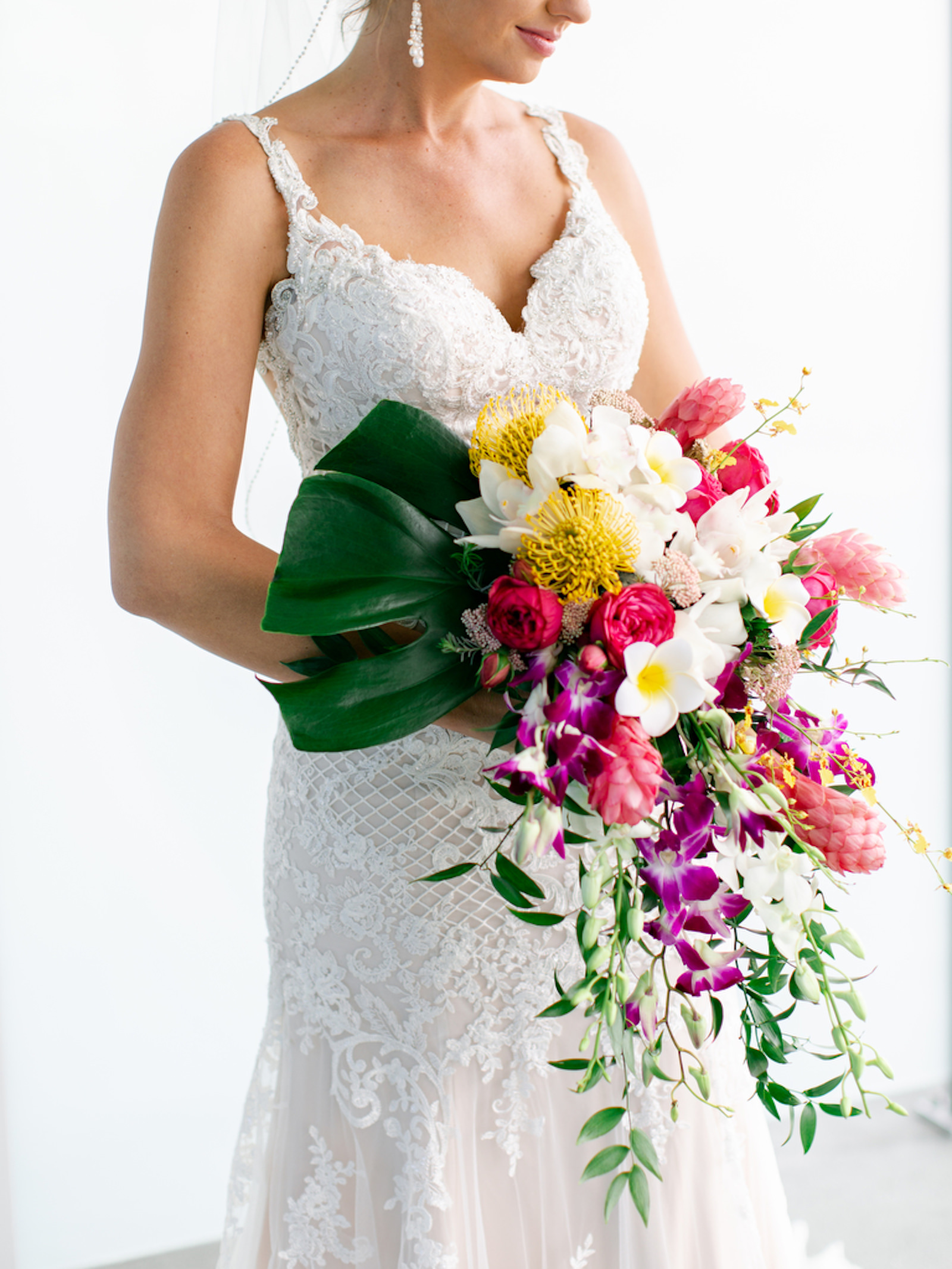 Tropical Elegant Bride in Delicate Lace Wedding Dress Holding Lush Colorful Floral Bouquet with Purple Orchids, Pink Roses, White, Yellow Pincushion Protea Flowers, Greenery and Palm Tree Leaf