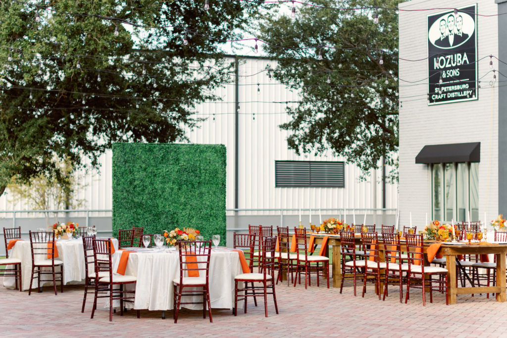 Boho Inspired Florida Wedding Reception and Decor, Autumn Wedding Theme with Warm Color Palette, Dark Red Chiavari Rental chairs, Round Tables with White Tablecloths and Burnt Orange Linens, Low Floral Centerpieces with Yellow Crystal Goblets, Greenery Boxwood Wall | Kozuba & Sons St. Petersburg Craft Distillery | Tampa Bay Wedding Planner Coastal Coordinating