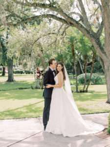 Outdoor Bride and Groom Portrait | V Back Embroidered Illusion Panel Allure Couture Designer Wedding Dress Bridal Gown with Long Cathedral Veil | Fall Wedding Bridal Bouquet with Red and White roses and Eucalyptus Greenery | Groom in Classic Black Suit Tux with Bow Tie