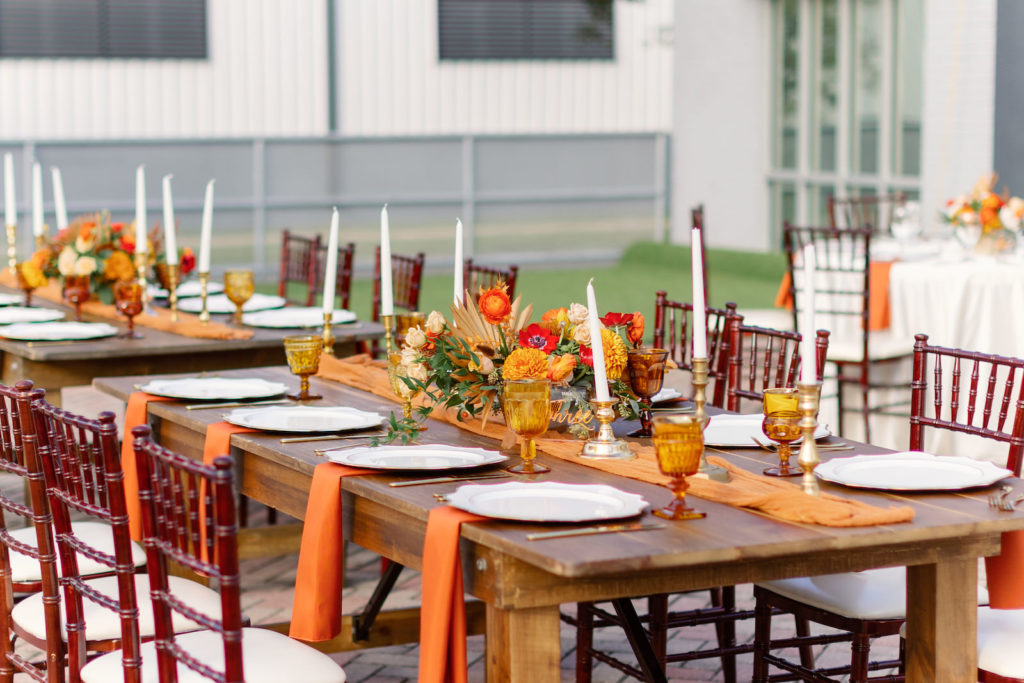 Modern Boho Inspired Florida Wedding Reception and Decor | Long Wooden Feasting Table with Burnt Orange Linen Table Runner with Long White Candles and Gold Candleholder, White Chargers, Orange, Golden Yellow and Red Floral Centerpiece | Tampa Bay Wedding Planner Coastal Coordinating