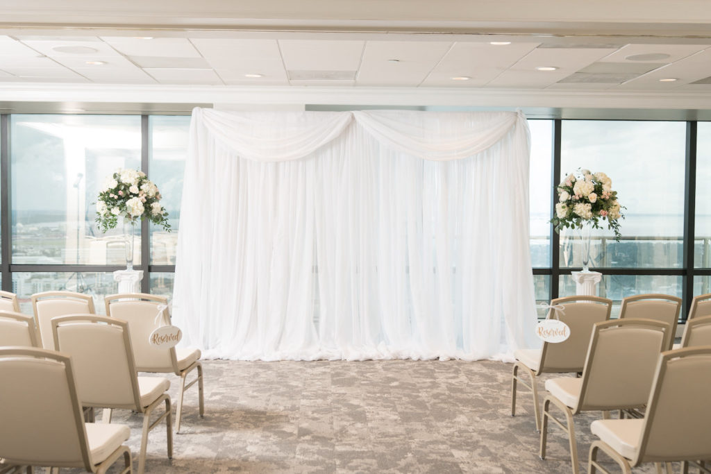 Indoor Wedding Ceremony at Downtown Tampa wedding Venue The Tampa Club | White Chiffon Pipe and Drape Ceremony Backdrop Background with Altar Floral Arrangements of White Hydrangea and White and Blush Pink Roses with Eucalyptus Greenery on Tall Clear Glass Vases | Carrie Wildes Photography