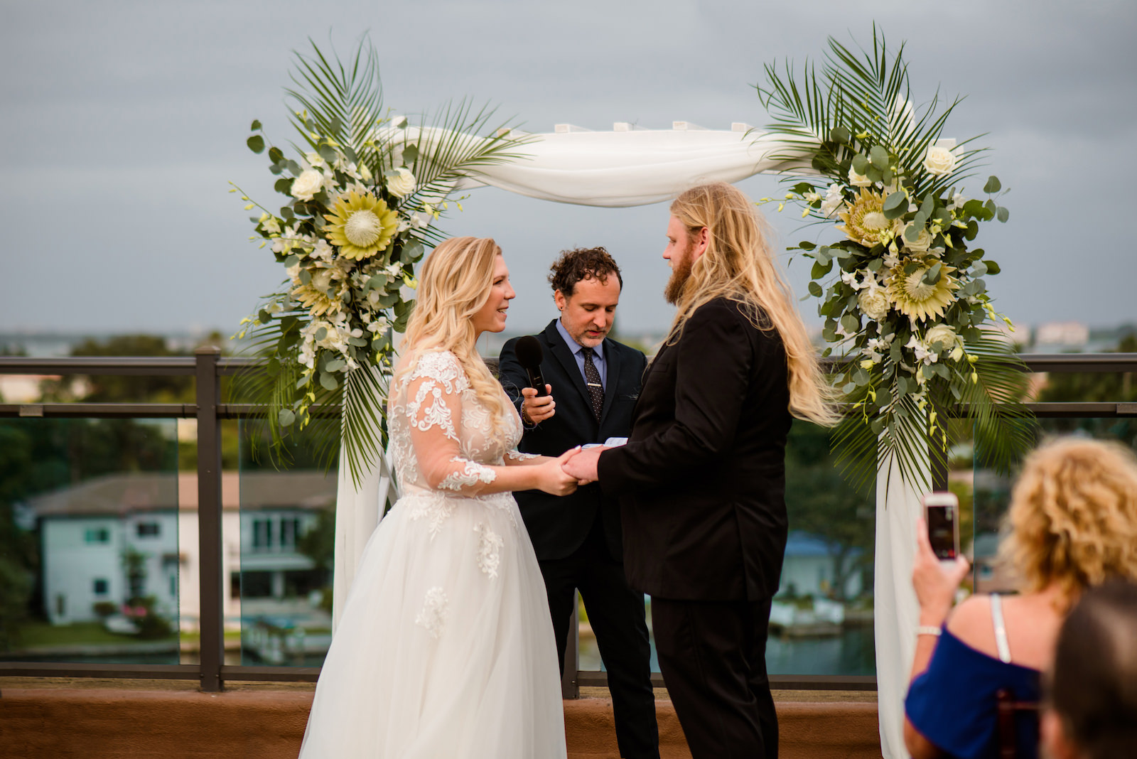 Florida Bride and Groom Exchanging Rooftop Wedding Ceremony Vows Under Arch with White Draping, Lush Neutral Tropical Boho Floral Arrangements, King Proteas, White Florals, Palm Fronds, Eucalyptus | Tampa Bay Wedding Planner Perfecting the Plan | Wedding Florist Iza's Flowers | St. Pete Waterfront Wedding Venue The Hotel Zamora