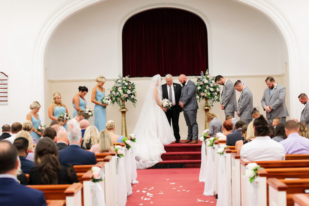 Traditional Tampa Church Wedding Ceremony with Wood Pews and White Ribbon Aisle Markers | Alter Floral Arrangements with Blush Pink and White Roses and Hydrangea with Eucalyptus Greenery on Gold Stands by Tampa Wedding Florist Monarch Events and Design | Groom and Groomsmen Wearing Classic Charcoal Grey Suits | Dusty Blue Periwinkle Bridesmaid Dresses