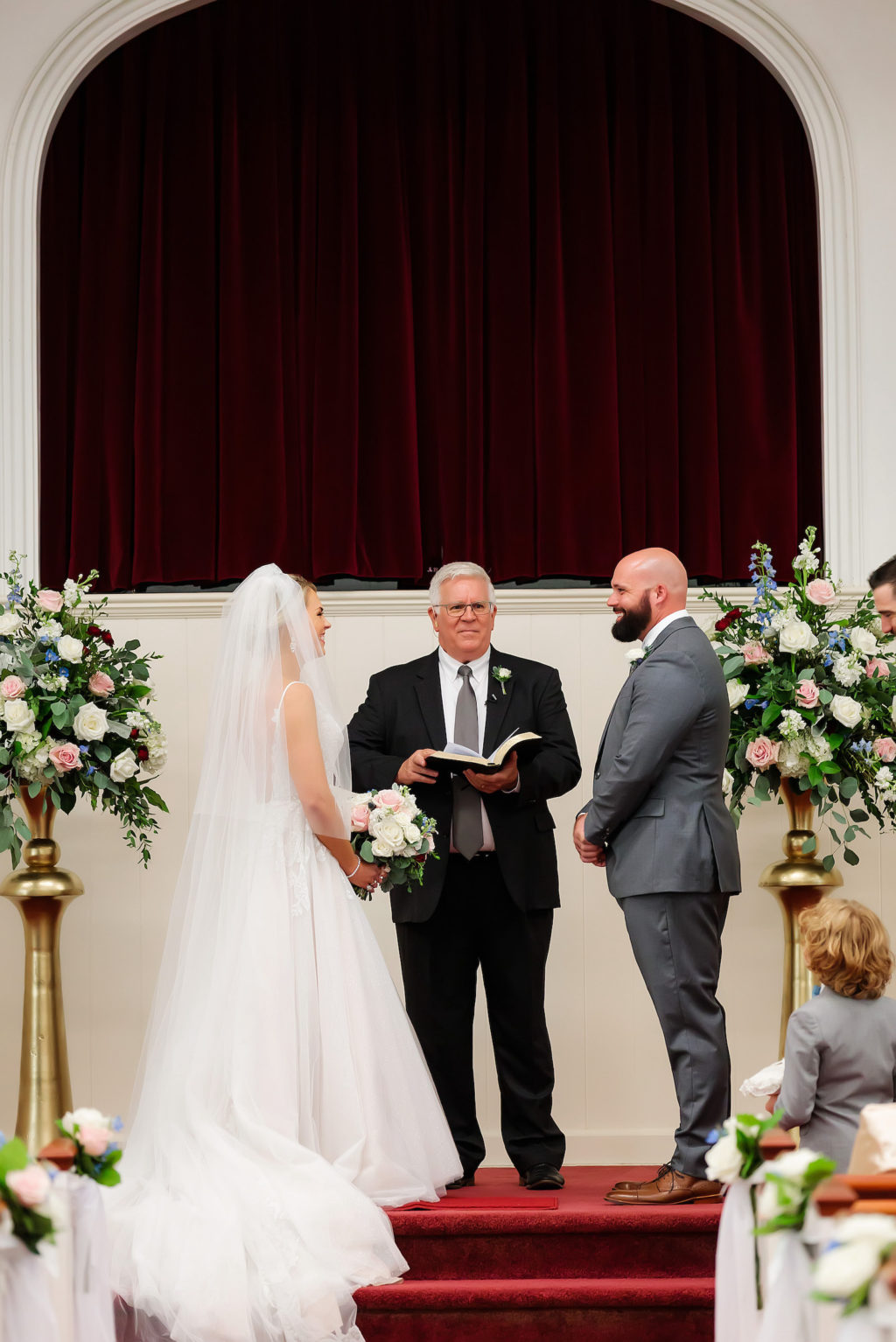Bride and Groom Exchanging Vows during Traditional Tampa Church Wedding Ceremony | Groom Wearing Charcoal Grey Suit | Alter Floral Arrangements with Blush Pink and White Roses and Hydrangea with Eucalyptus Greenery on Gold Stands by Tampa Wedding Florist Monarch Events and Design