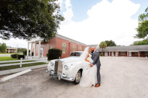 Outdoor Bride and Groom Portrait in front of White Classic Car Rolls Royce at Traditional Church Ceremony | Long Cathedral Veil | A Line Ballgown Tulle V Neck Lace Wedding Dress by Hayley Paige Designer | Groom Wearing Classic Charcoal Grey Suit