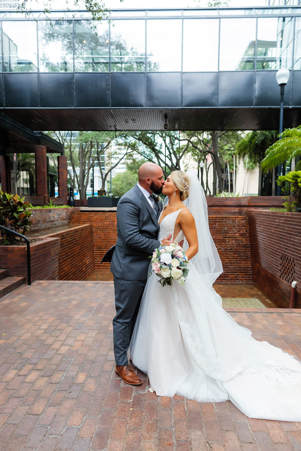 Outdoor Bride and Groom Portrait at Tampa Wedding Venue Hilton Tampa Downtown | Long Cathedral Veil | A Line Ballgown Tulle V Neck Lace Wedding Dress by Hayley Paige Designer | Groom Wearing Classic Charcoal Grey Suit