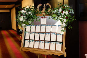 Wedding Seating Chart Mirror with Ornate Gold Frame and Alphabetized Seating Arrangement Cards with Blue and Cranberry Satin Ribbons | Floral Garland Spray Arrangement with White and Maroon Roses and Eucalyptus Greenery by Tampa Wedding Florist Monarch Events and Design