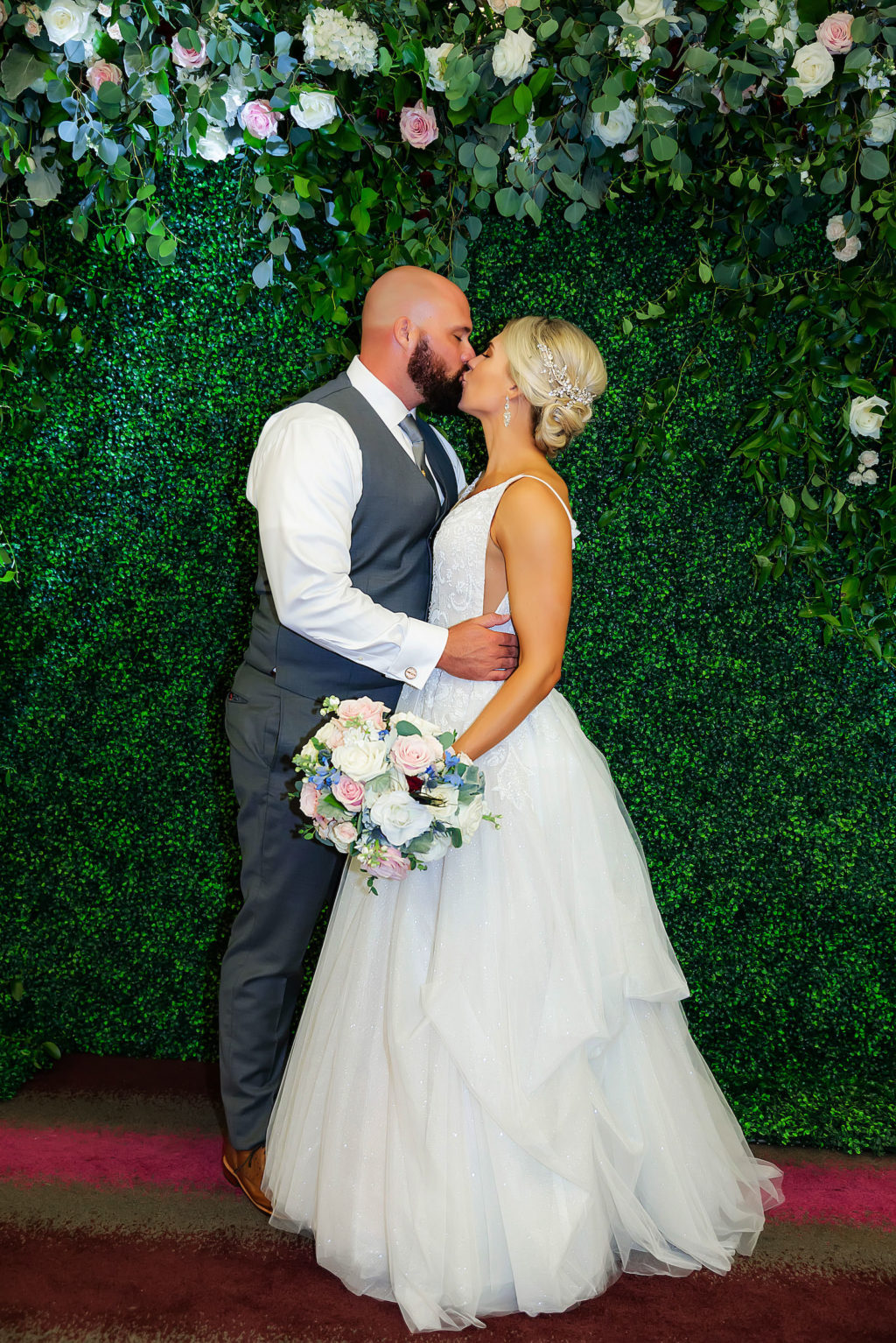 Indoor Bride and Groom Portrait in front of Boxwood Hedge Greenery Wall with Blush Pink and White Roses and Dusty Blue Eucalyptus Greenery by Tampa Wedding Florist Monarch Events and Design | A Line Ballgown Tulle V Neck Lace Wedding Dress by Hayley Paige Designer | Groom Wearing Classic Charcoal Grey Suit