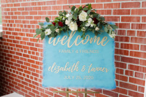 Acrylic Wedding Welcome Sign with Dusty Blue and Gold Calligraphy and Floral Spray Arrangement of White Roses and Eucalyptus Greenery