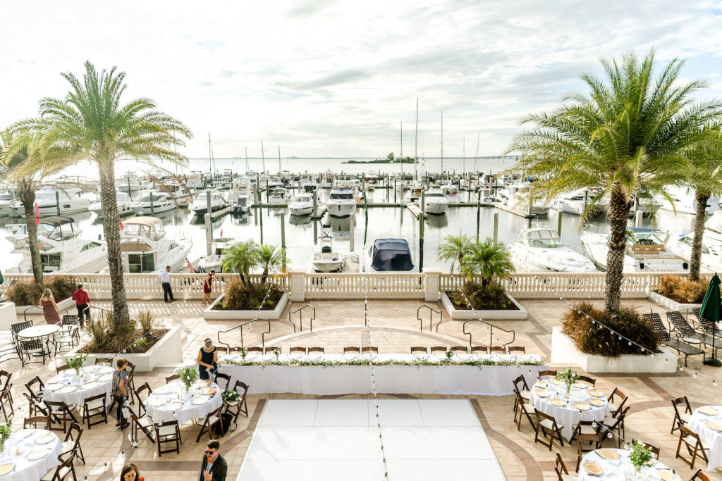 Outdoor wedding Reception at Tampa wedding Venue westshore Yacht Club | Round tables with white linens and mahogany wood folding chairs with gold charger plates and greenery centerpieces | long wedding head table with white dance floor