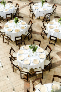 Outdoor wedding Reception with Round tables with white linens and mahogany wood folding chairs with gold charger plates and greenery centerpieces