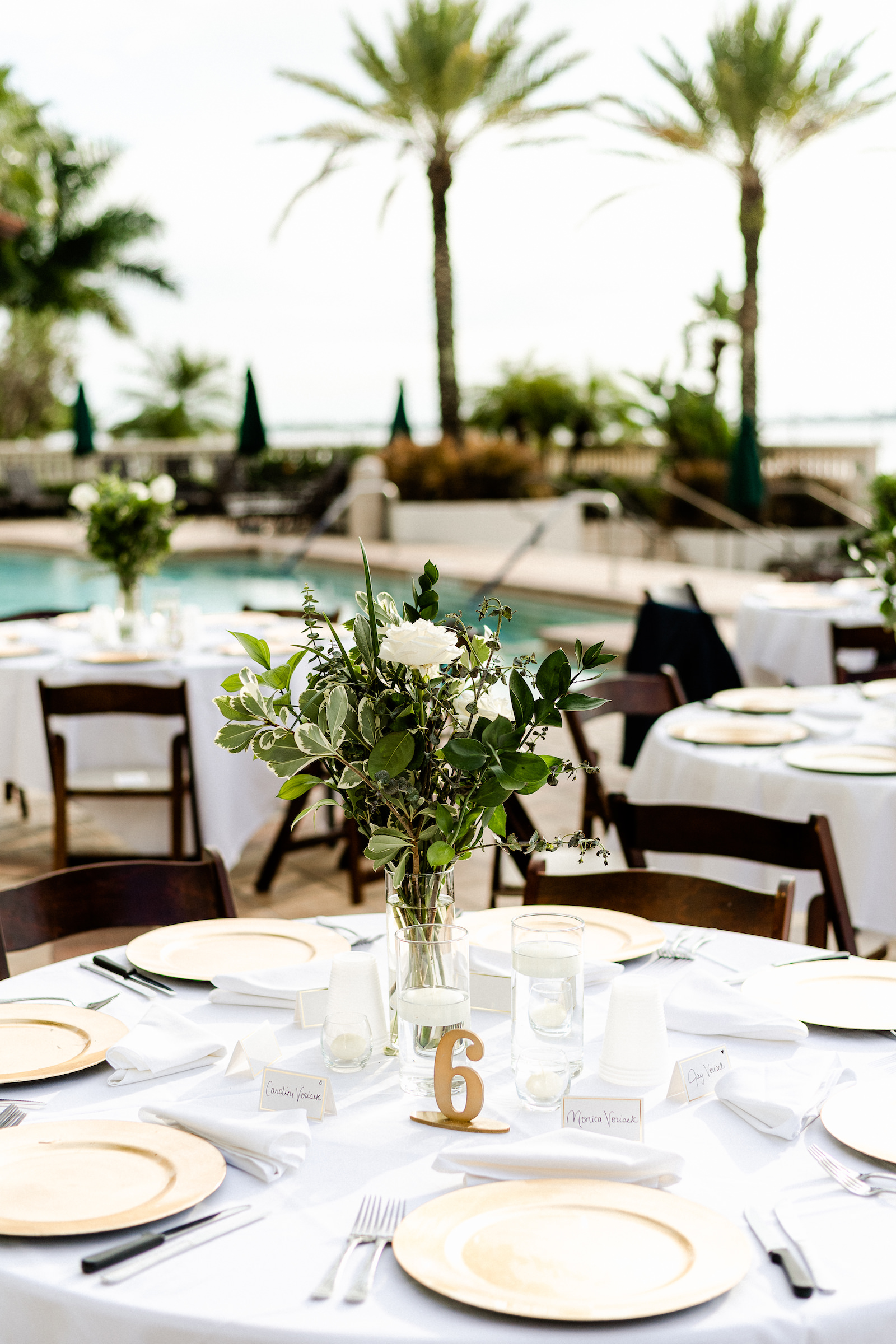 Outdoor wedding Reception with Round tables with white linens and mahogany wood folding chairs with gold charger plates and greenery centerpieces | Westshore Yacht Club | Dewitt for Love Photography