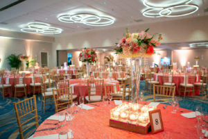 Tampa Clearwater Beach Indian Wedding at Hilton Clearwater Beach Wedding Venue | Ballroom Hotel Wedding Reception Tables with Gold Chiavari Chairs and Coral Pink Table Cloth Linens and White Napkins | Tall Floral Centerpieces with Pink Peach Coral Roses and White Hydrangea surrounded by Floating Candles | Hilton Clearwater Beach