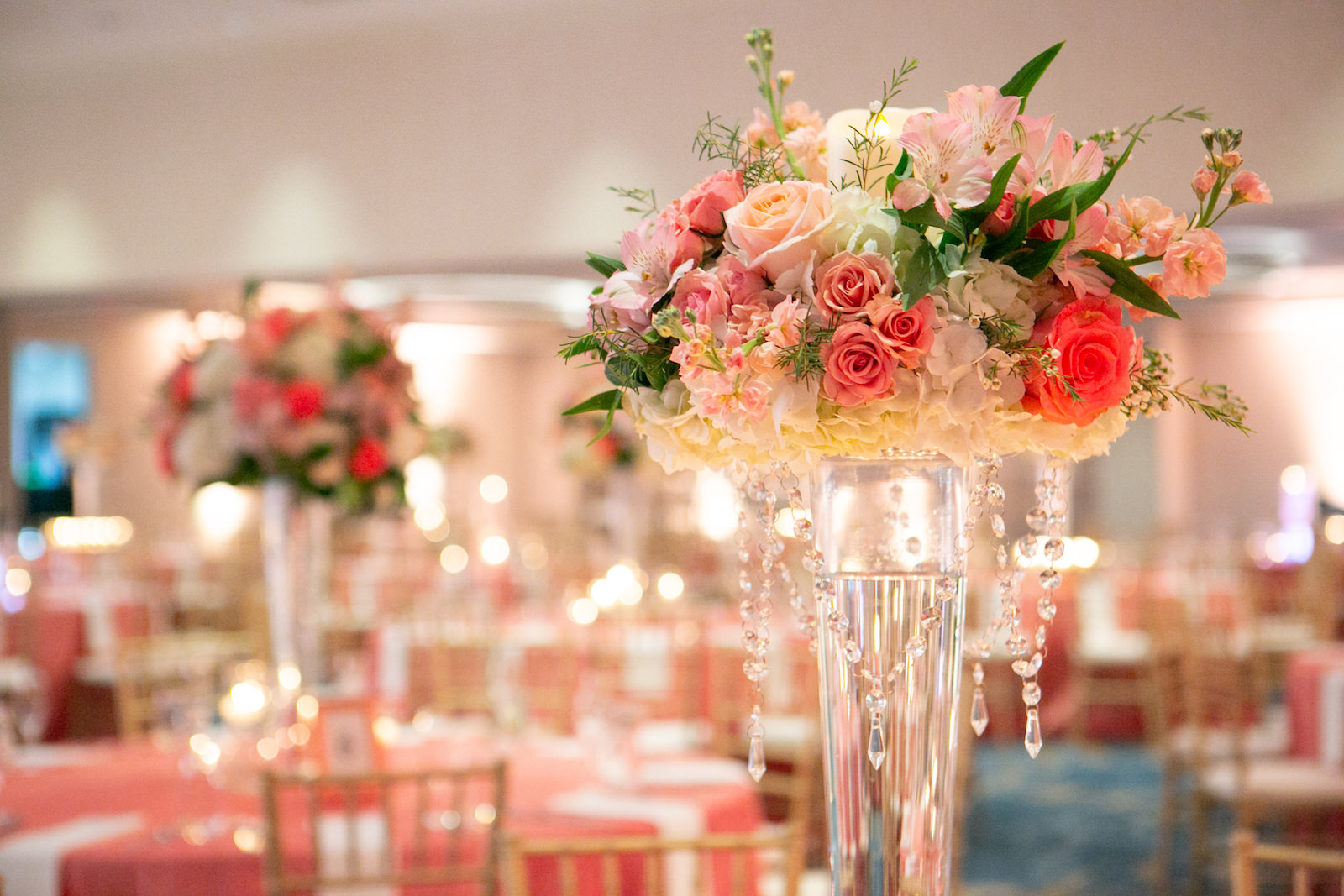 Tampa Clearwater Beach Indian Wedding at Hilton Clearwater Beach Wedding Venue | Ballroom Hotel Wedding Reception Tables with Gold Chiavari Chairs and Coral Pink Table Cloth Linens and White Napkins | Tall Floral Centerpieces with Pink Peach Coral Roses and White Hydrangea surrounded by Floating Candles