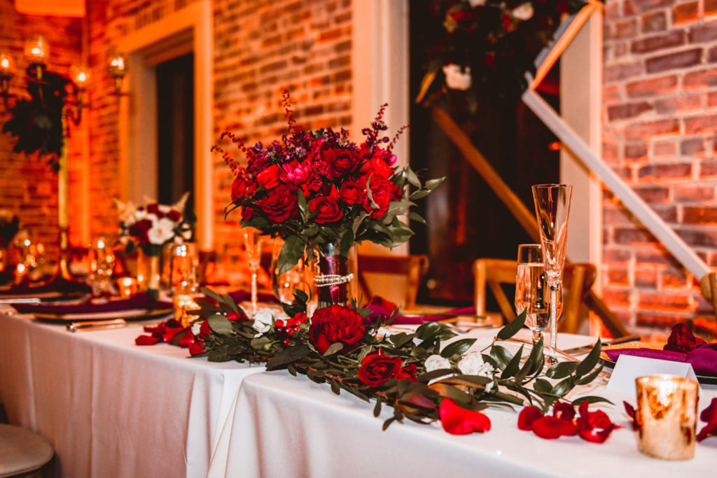 Botanical Inspired Wedding Reception Decor, Long Feasting Sweetheart Table with Greenery, Red Roses, Wooden Cross Back Chairs, Geometric Sculpture and Red Brick Backdrop | Tampa Bay Wedding Planner Special Moments Event Planning | Downtown St. Pete Wedding Venue Red Mesa Events