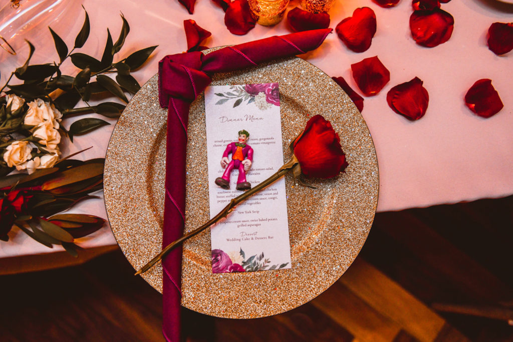 Botanical Inspired Wedding Reception Decor, Gold Glitter Charger with Burgundy Red Linen Napkin, Red Roses and Joker Figurine | Tampa Bay Wedding Planner Special Moments Event Planning