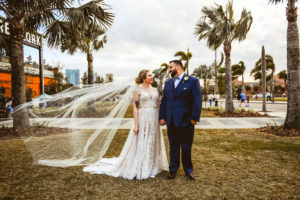 Florida Bride in Lace and Tulle Wedding Dress with Veil Blowing in the Wind, Groom in Blue Tuxedo Outside Tampa Industrial Wedding Venue Armature Works