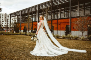 Florida Bride in Lace and Tulle Boho Wedding Dress with Neutral Ribbon Belt, Full Length Veil Holding White and Greenery Floral Bouquet | Tampa Industrial Wedding Venue Armature Works