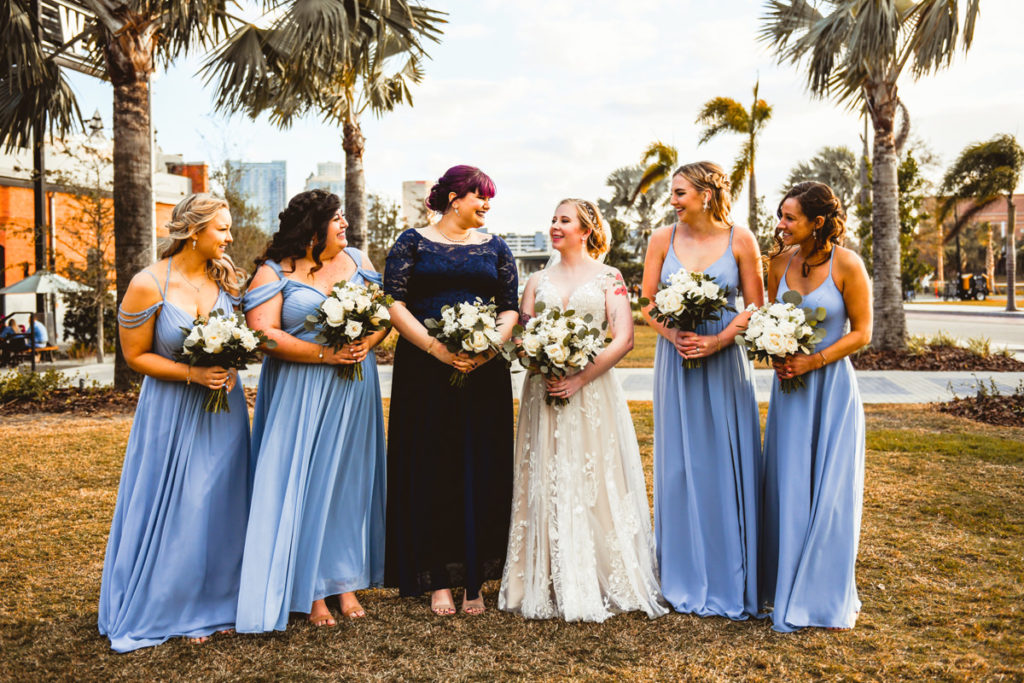 Florida Bride with Bridesmaids in Mix and Match Dusty Blue Dresses Holding White and Greenery Floral Bouquets | Tampa Industrial Wedding Venue Armature Works | Tampa Bay Wedding Hair and Makeup Femme Akoi