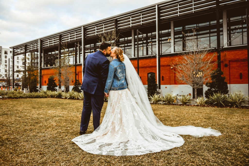 Boho Bride in Lace and Tulle Wedding Dress and Custom Denim Jacket with Full Length Veil Kissing Groom Outside Tampa Industrial Wedding Venue Armature Works