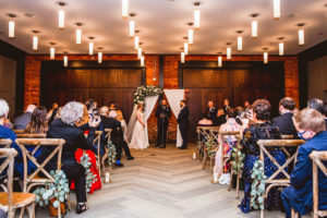 Industrial Chic Wedding Ceremony, Bride and Groom Exchangin Wedding Vows, Wooden Arch with White Linen Drapery, Lush Greenery and Ivory Floral Arrangement, Wooden Cross Back Chairs with Eucalyptus Hanging | Tampa Wedding Venue Armature Works