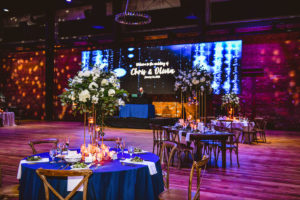 Industrial Elegant Wedding Reception Decor, Purple Uplighting, Round Tables and Long Feasting Wooden Tables, Wooden Cross Back Chairs, Tall Ivory and Greenery Floral Centerpieces | Tampa Wedding Venue Armature Works