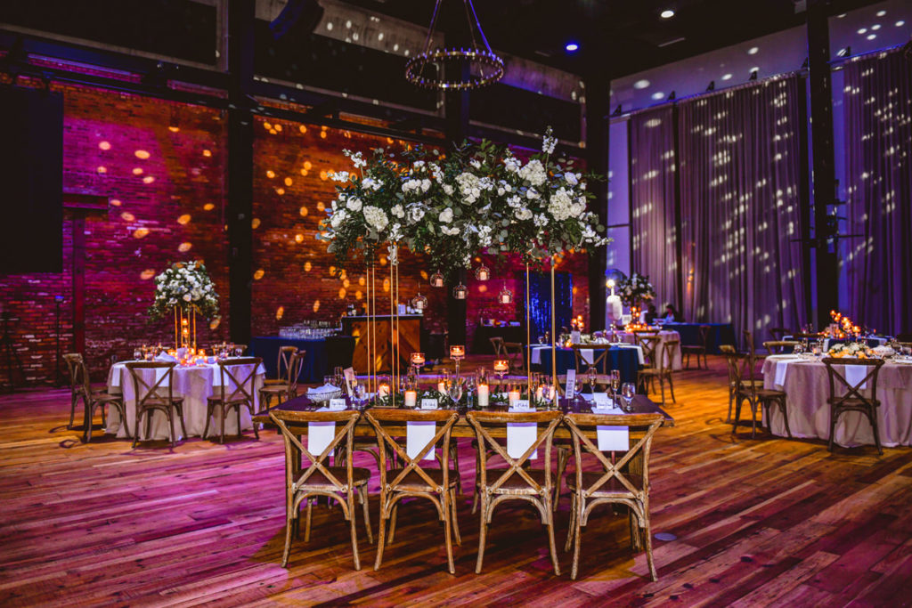 Industrial Elegant Wedding Reception Decor, Purple Uplighting with Yellow Dot Projections, Round Tables and Long Feasting Wooden Tables, Wooden Cross Back Chairs, Tall Ivory and Greenery Floral Centerpieces | Tampa Wedding Venue Armature Works