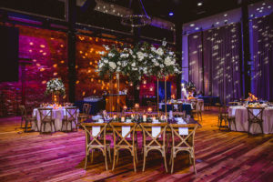 Industrial Elegant Wedding Reception Decor, Purple Uplighting with Yellow Dot Projections, Round Tables and Long Feasting Wooden Tables, Wooden Cross Back Chairs, Tall Ivory and Greenery Floral Centerpieces | Tampa Wedding Venue Armature Works