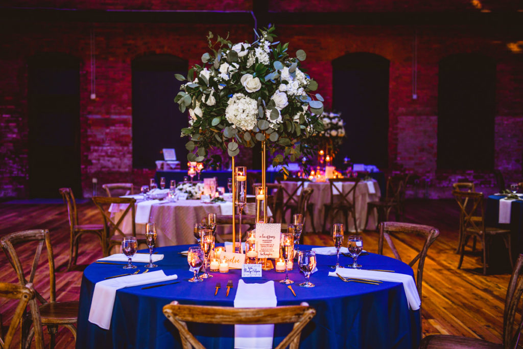 Industrial Chic Wedding Reception Decor, Round Table with Blue Tablecloth, White Linen Napkins, Tall Gold Stand with Eucalyptus, White Roses and Hydrangeas, Candles | Tampa Wedding Venue Armature Works