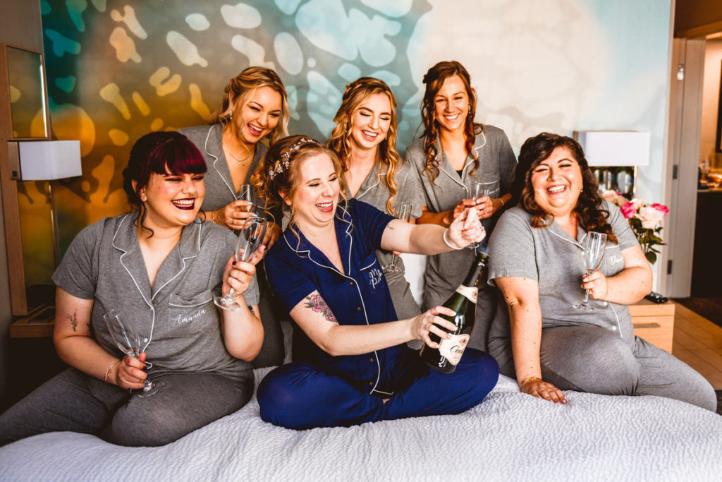 Florida Bride in Blue Pajamas and Bridesmaids in Matching Gray Pajamas Popping Bottle of Champagne | Tampa Bay Wedding Hair and Makeup Femme Akoi