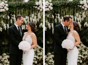 Outdoor Bride and Groom Portrait in front of Wedding Ceremony Backdrop wood Arch Arbor with Greenery and White Roses and Suspended Hanging Stock Flowers | Groom Wearing Classic Black Suit Tux | White Peony Bridal Bouquet | Sheath Illusion Lace Bridal Gown wedding Dress