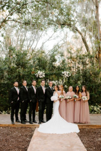 Outdoor Wedding Party Portrait in front of Wedding Ceremony Backdrop wood Arch Arbor with Greenery and White Roses and Suspended Hanging Stock Flowers | Groom and Groomsmen Wearing Classic Black Suit Tux | White Peony Bridal Bouquet | Sheath Illusion Lace Bridal Gown wedding Dress | Long Dusty Rose Mauve Pink Bridesmaid Dresses