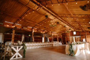 Wood Barn Reception at Plant City Wedding Venue Florida Rustic Barn Weddings | Long Feasting Tables with White Linens and Eucalyptus Greenery Garland Centerpieces and Wood Cross Back Chairs under a Canopy of String Lights